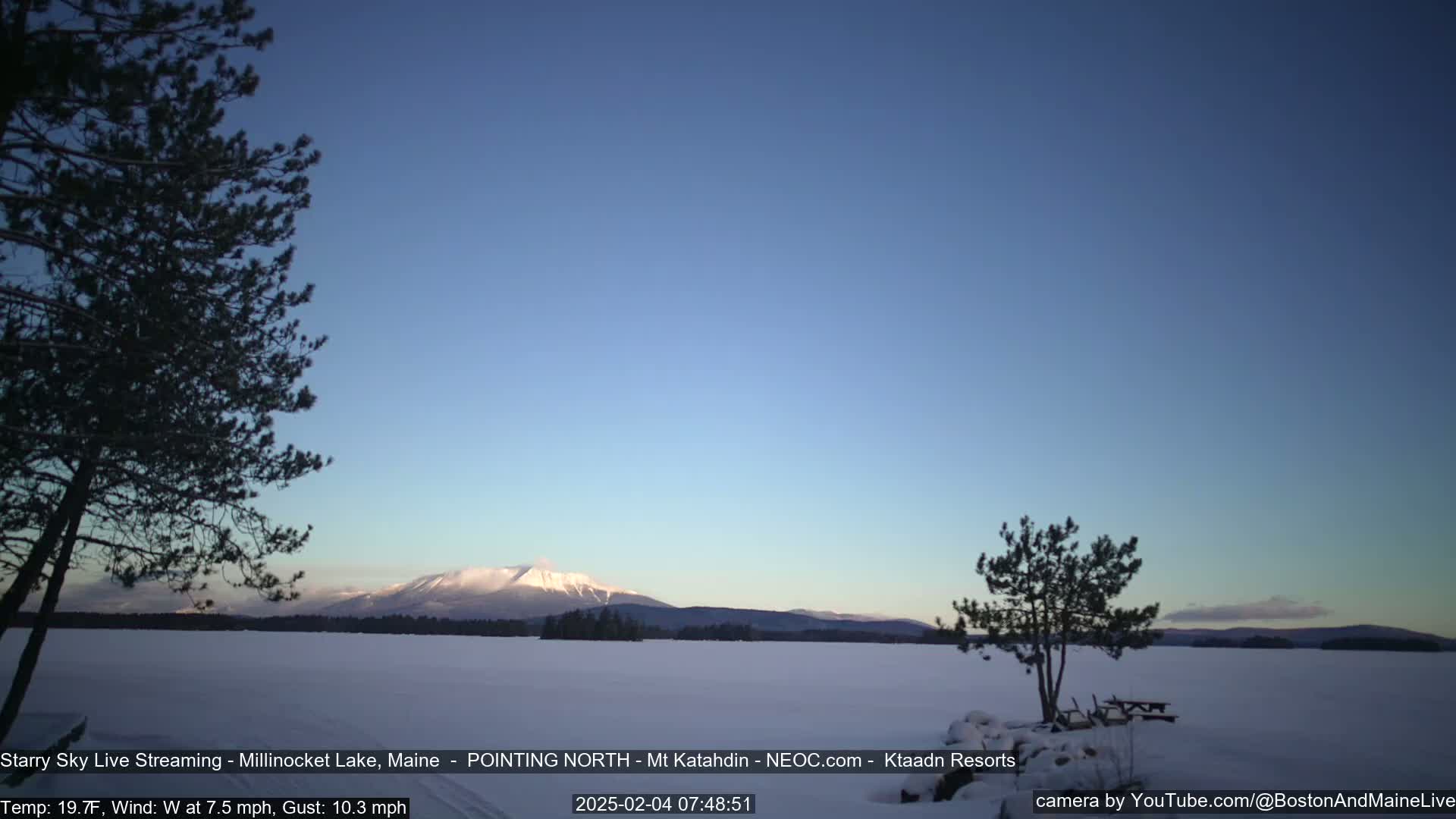 A snow-covered lake stretches to a distant mountain range under a clear, twilight sky with a light westerly wind.