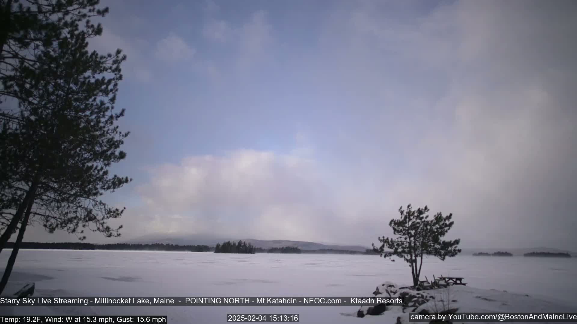A snow-covered lake stretches to a distant mountain range under a mostly cloudy, light-blue sky.
