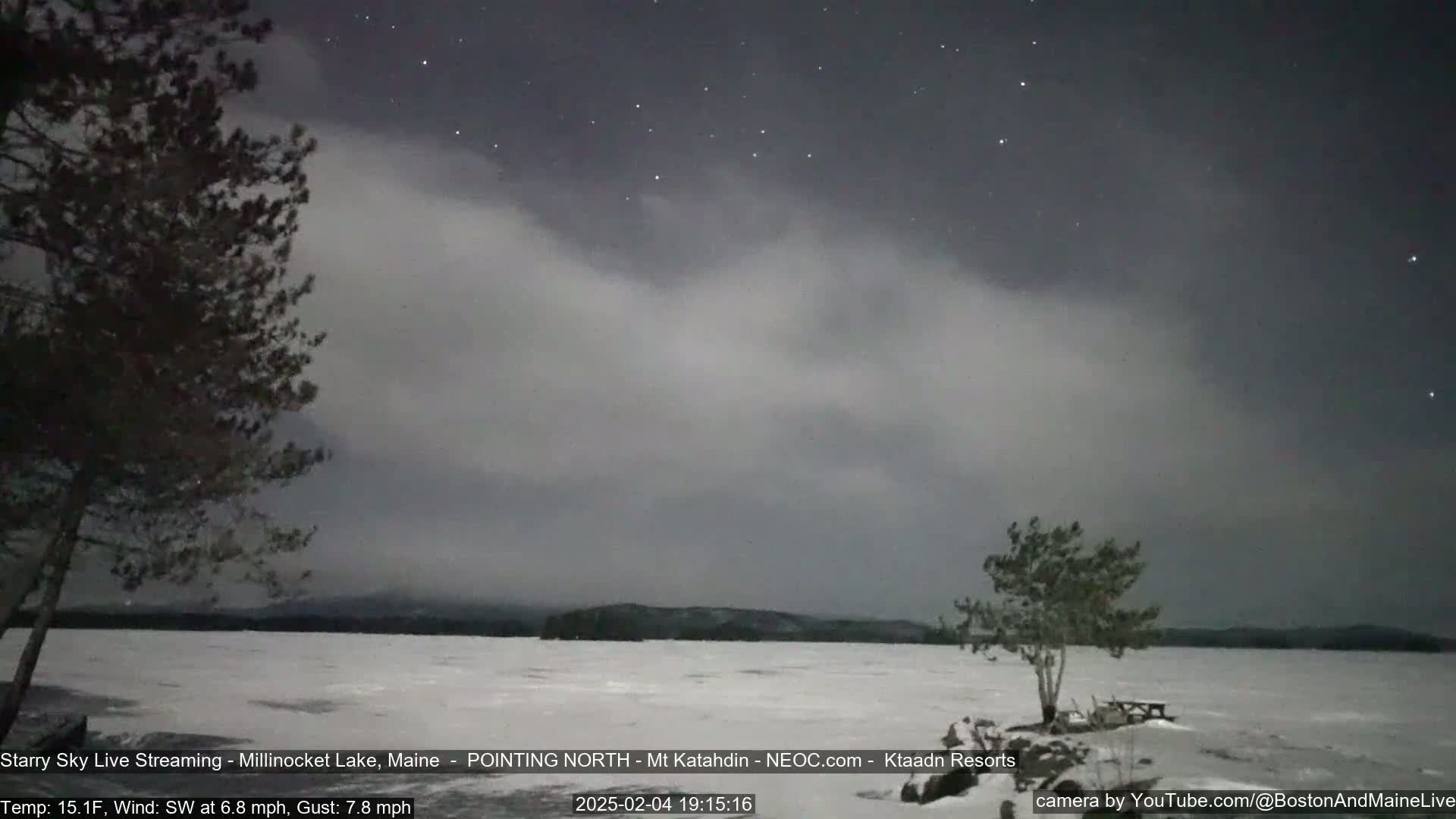 A snow-covered lake is visible under a partly cloudy night sky dotted with stars, with a few trees in the foreground.