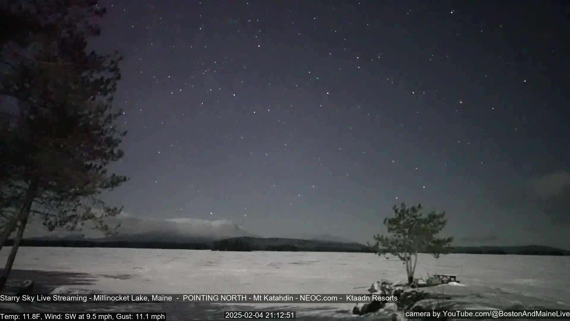 A snow-covered lake is visible under a starry night sky, with a few trees and a mountain range in the distance; the weather is cold, with a southwest wind.