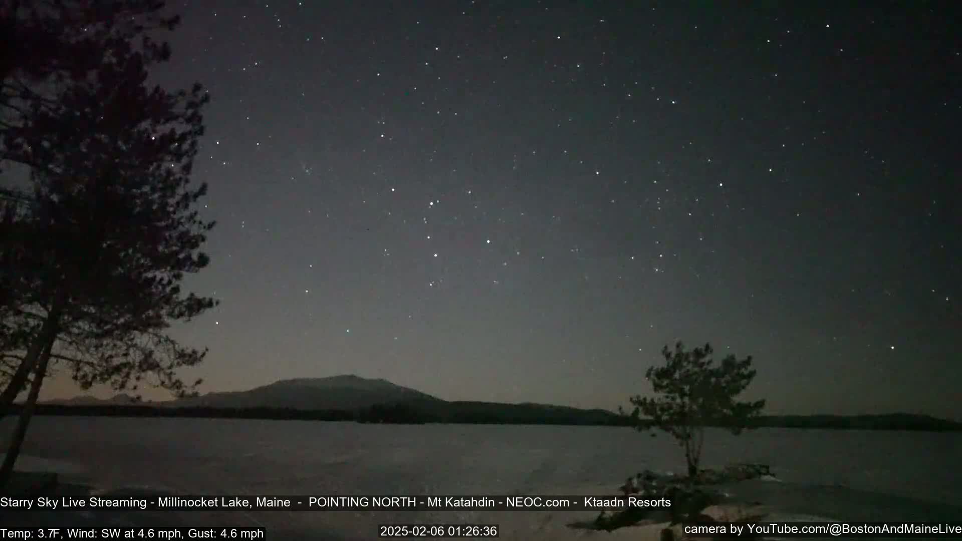 A starry night sky over a calm lake, with silhouetted trees in the foreground and mountains in the distance, under a slight southwest wind.