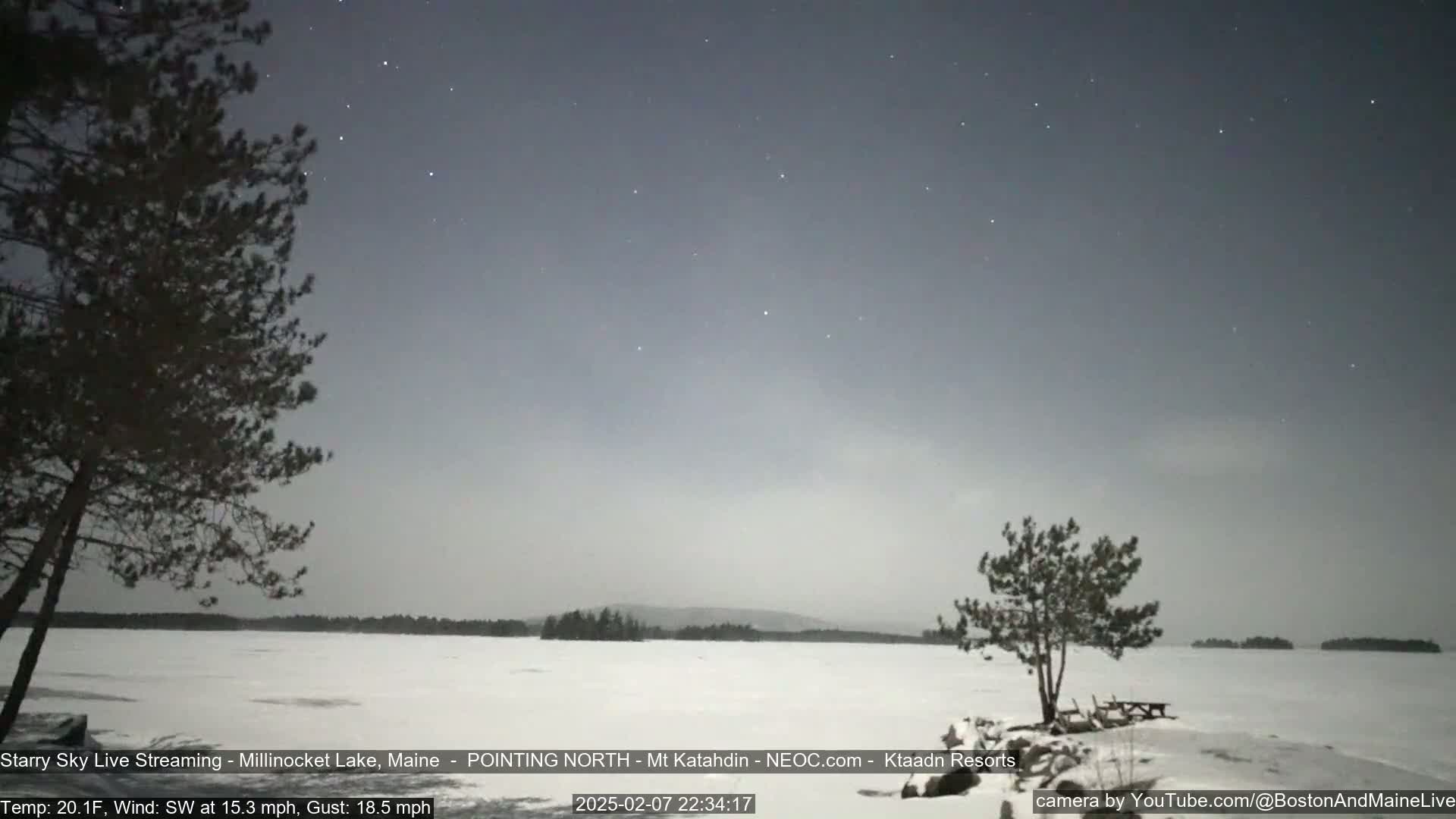 A snow-covered lake is visible under a mostly clear night sky dotted with stars, with a few trees and a picnic table in the foreground, and a light wind blowing.