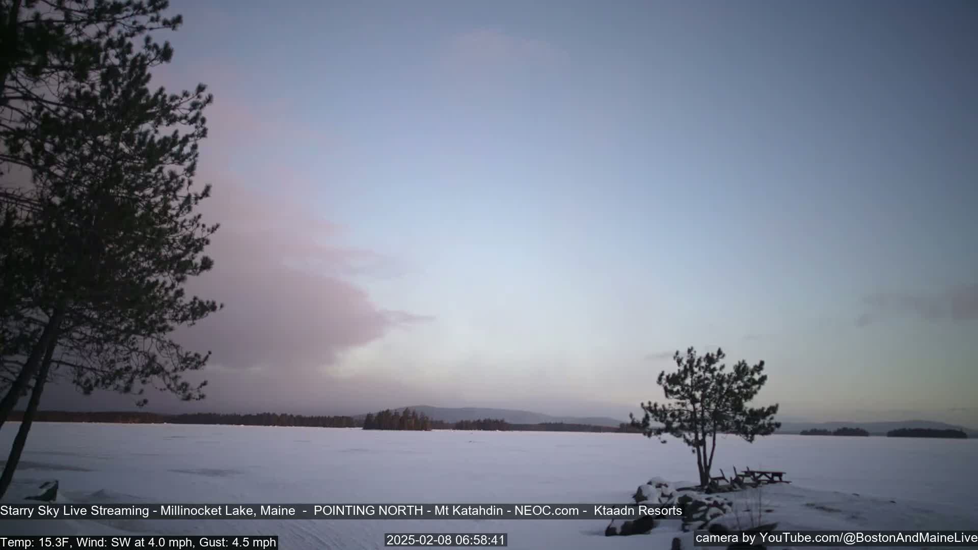 A snow-covered lake is seen at dawn under a mostly clear, light-blue sky with a few clouds, framed by dark evergreen trees on either side.