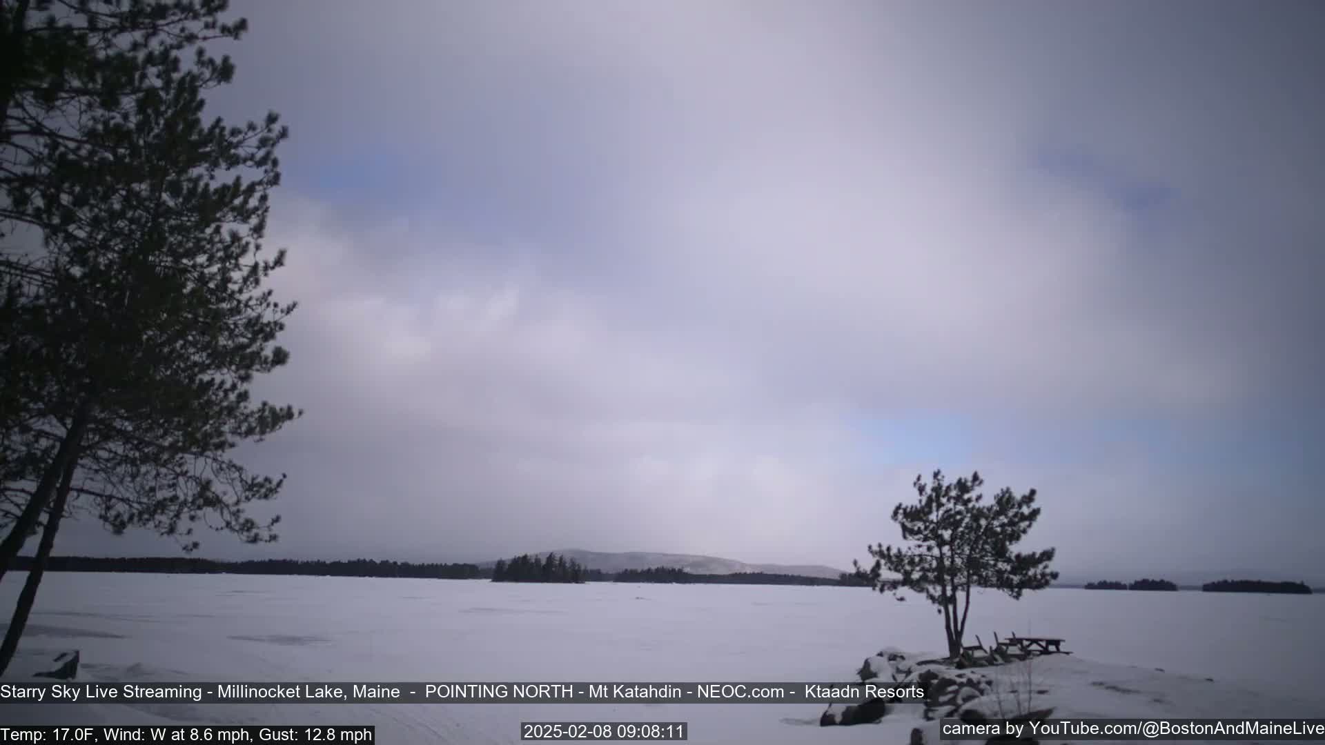 A snow-covered lake stretches to a distant shoreline under a mostly cloudy, light sky.