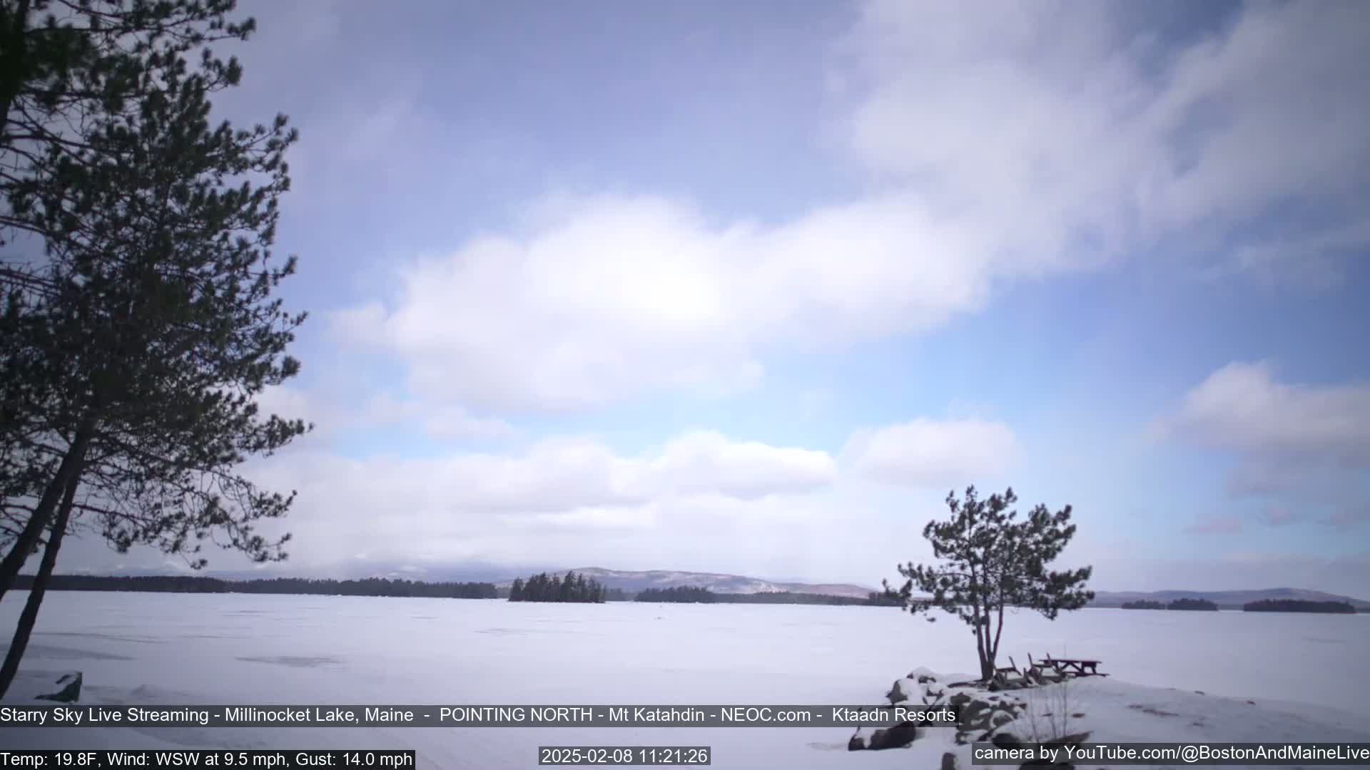 A mostly sunny, windy winter day with a snow-covered frozen lake, sparse trees, and distant mountains.