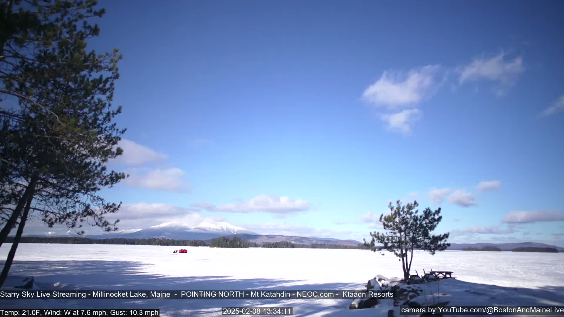 A snow-covered lake stretches to a mountain range under a mostly clear blue sky with a few scattered clouds; a small red structure is visible on the ice.