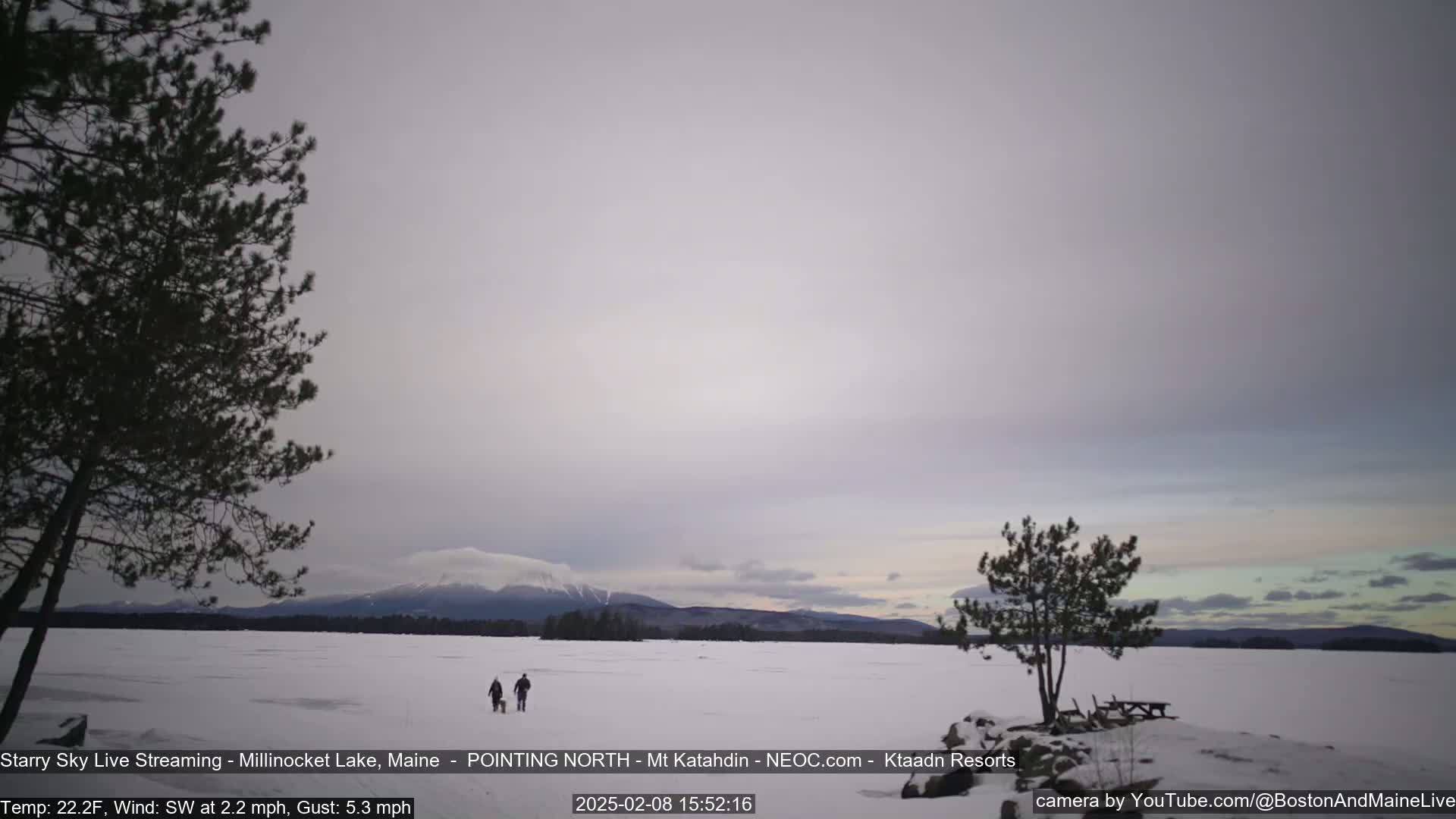 Two people walk a dog across a snow-covered frozen lake under a mostly cloudy sky with mountains in the distance.