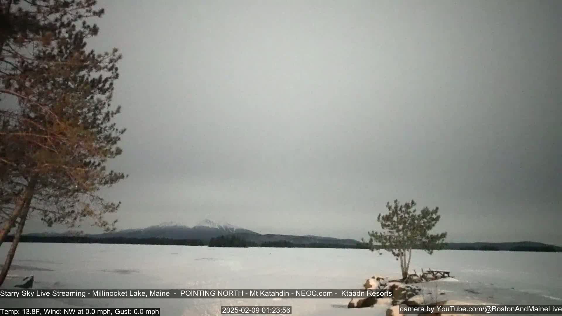 A snow-covered lake is shown under an overcast sky, with trees in the foreground and mountains in the distance.