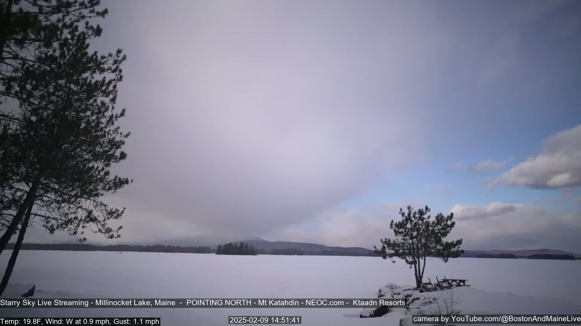 A snow-covered lake is seen under a mostly cloudy sky, with a few trees and distant mountains visible.