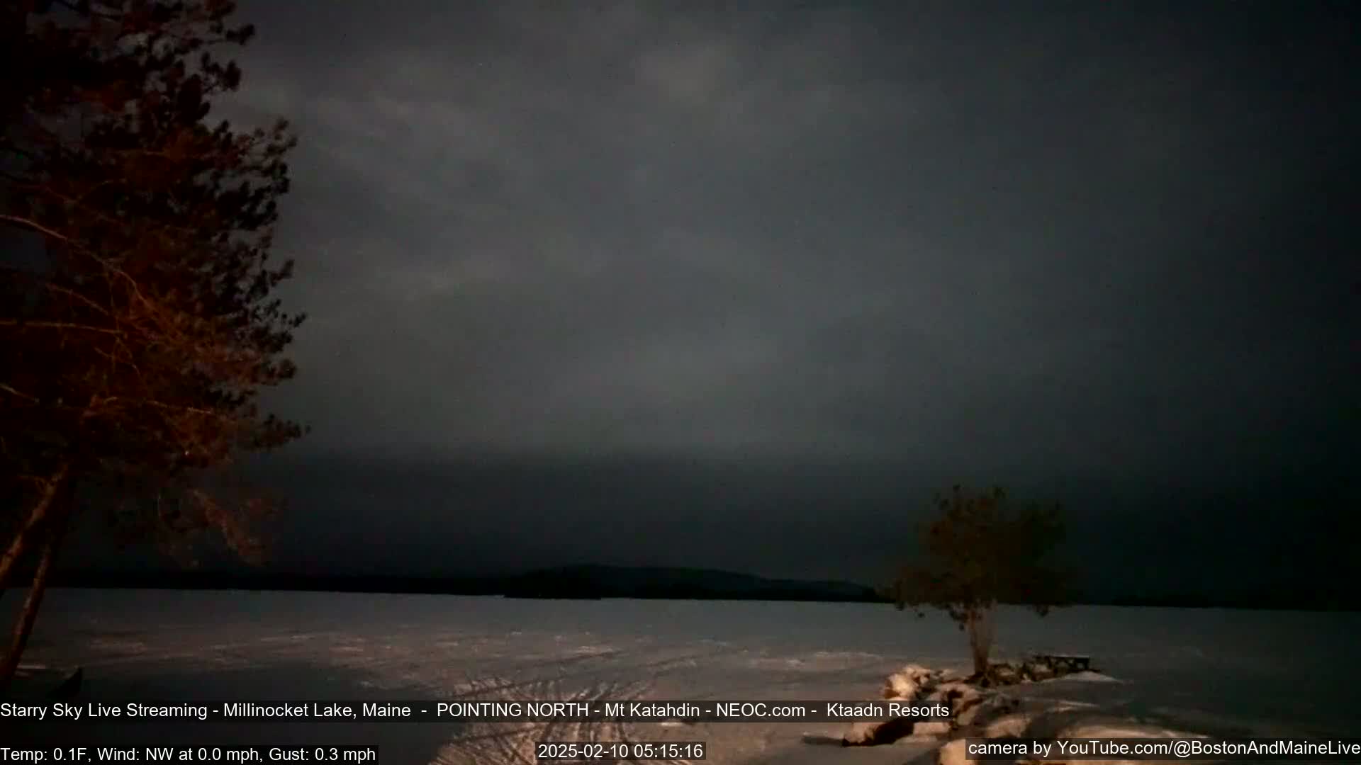 A dark, still nighttime scene shows a frozen lake under a mostly cloudy sky, with two silhouetted trees in the foreground.