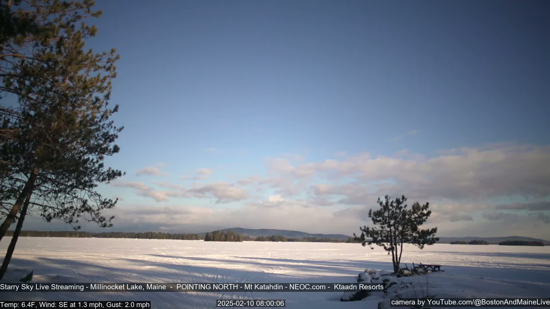 A snow-covered lake stretches to a treeline under a mostly clear, light-blue sky with a few clouds.