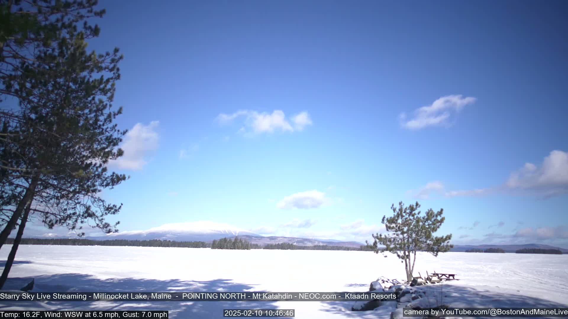 A snow-covered lake stretches to a distant mountain range under a mostly clear blue sky with a few scattered clouds.