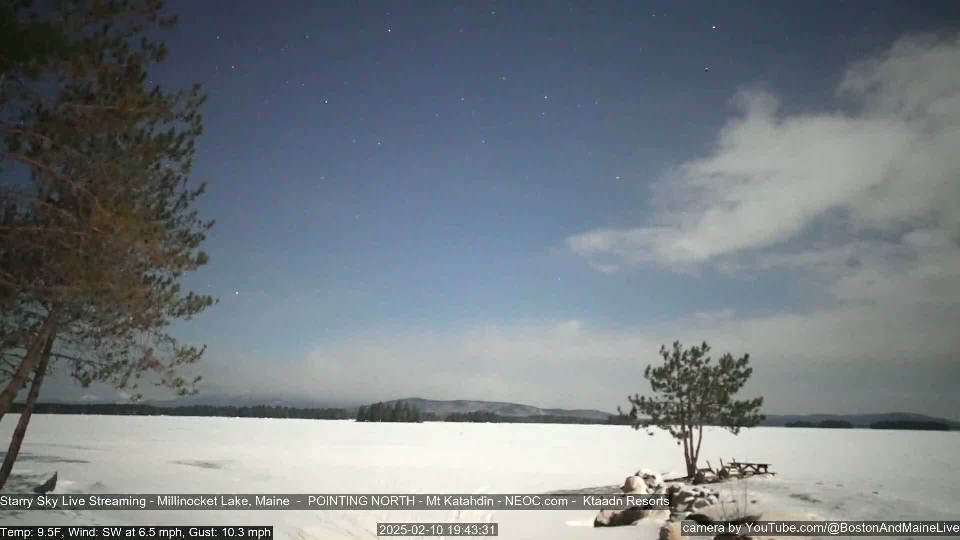 Under a partly cloudy, starry night sky, a snow-covered lake stretches to a dark treeline, with a small tree and picnic table visible in the foreground.
