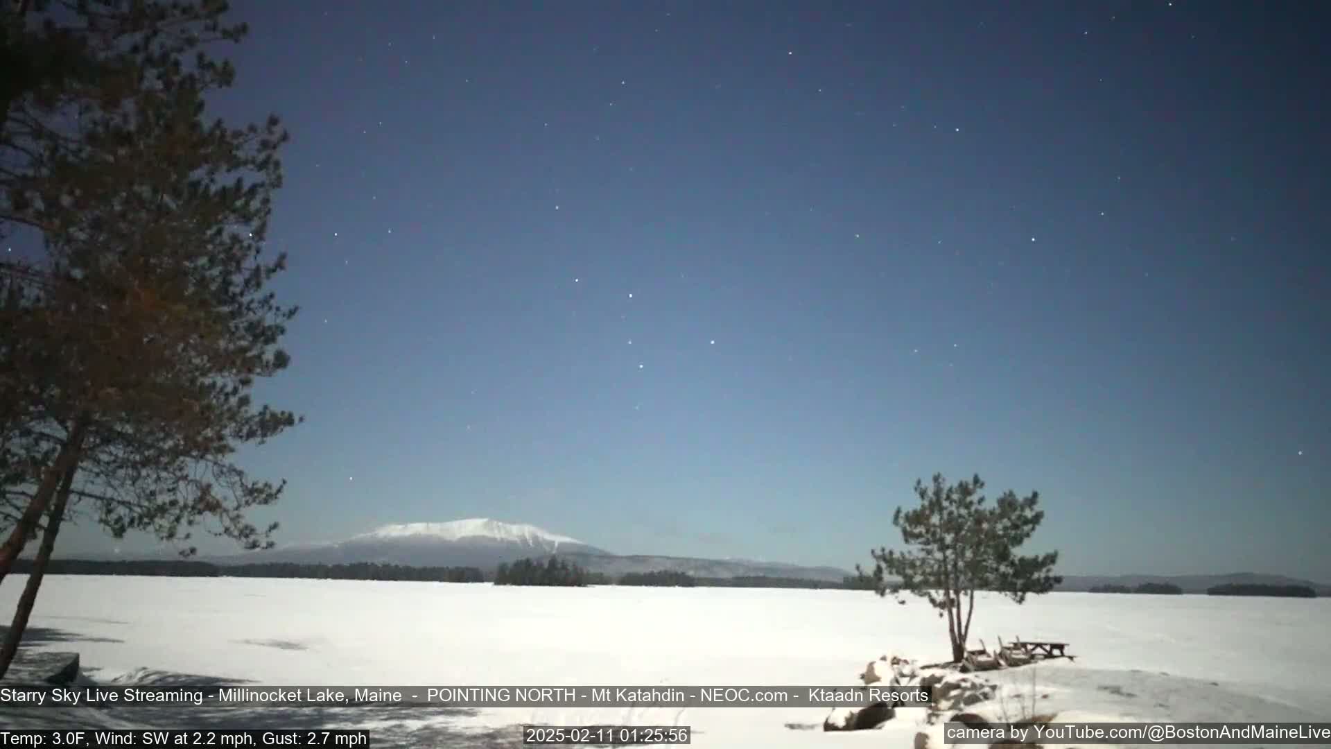 A starry night sky over a snow-covered frozen lake with a distant mountain range under a clear, dark sky.