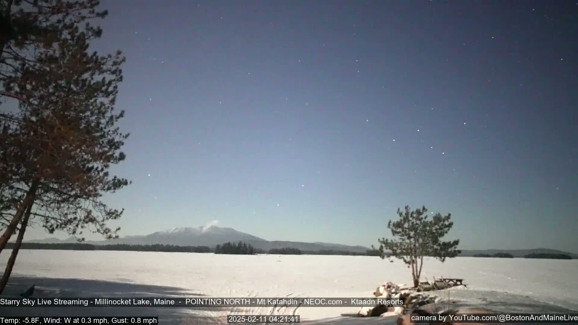 A snow-covered lake is seen under a starry night sky with a mountain range in the distance, and a slight wind.