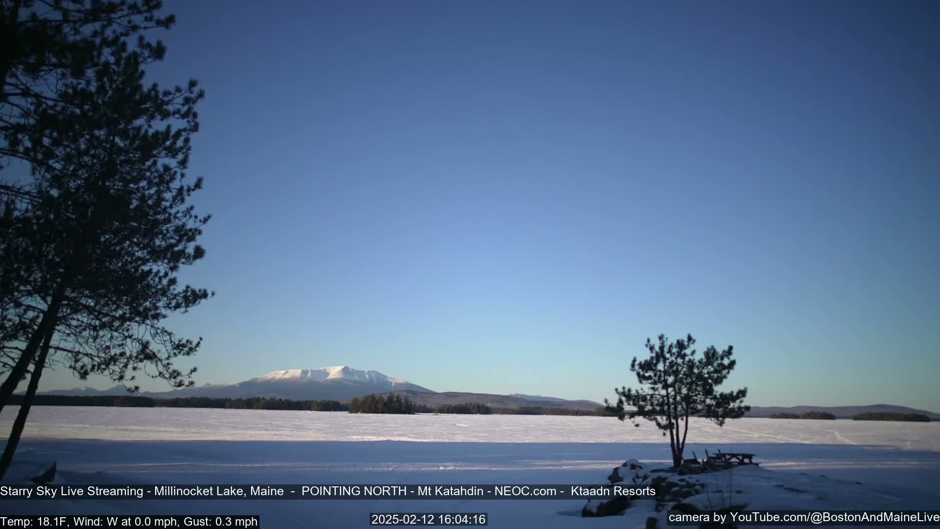A snow-covered lake stretches out to a distant, snow-capped mountain range under a clear, blue sky.
