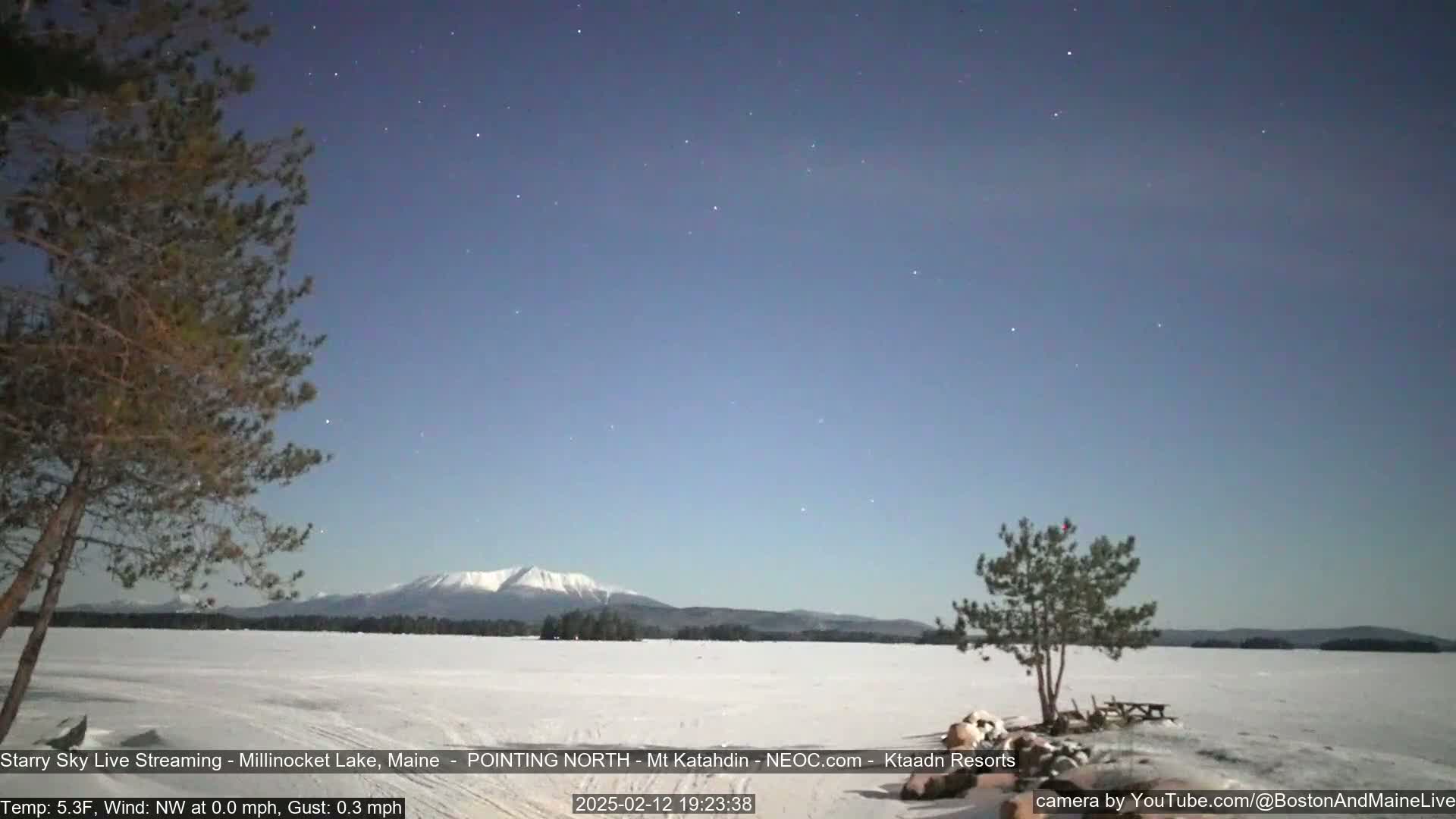 A snow-covered lake stretches to a mountain range under a clear, starry night sky.
