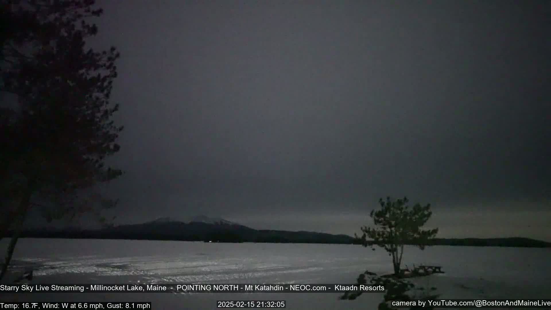 A dark, overcast sky hangs over a snow-covered lake with trees in the foreground and distant mountains visible on the horizon.
