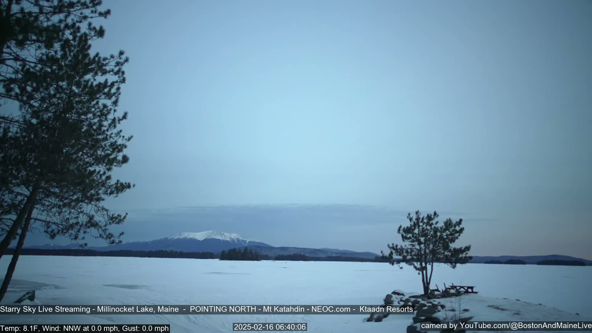 A snow-covered lake stretches towards a mountain range under a pale, twilight sky.
