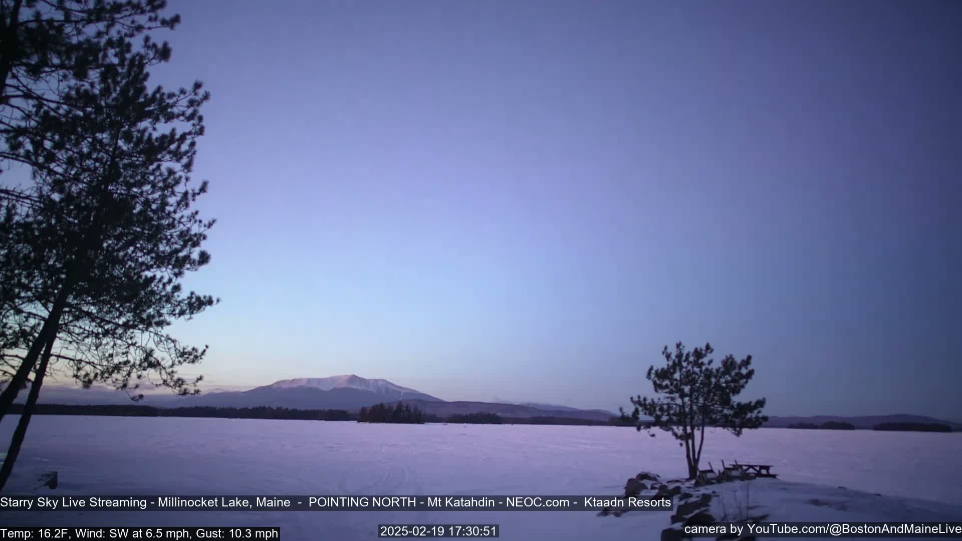A snow-covered lake stretches towards distant mountains under a twilight sky, with two silhouetted trees in the foreground.
