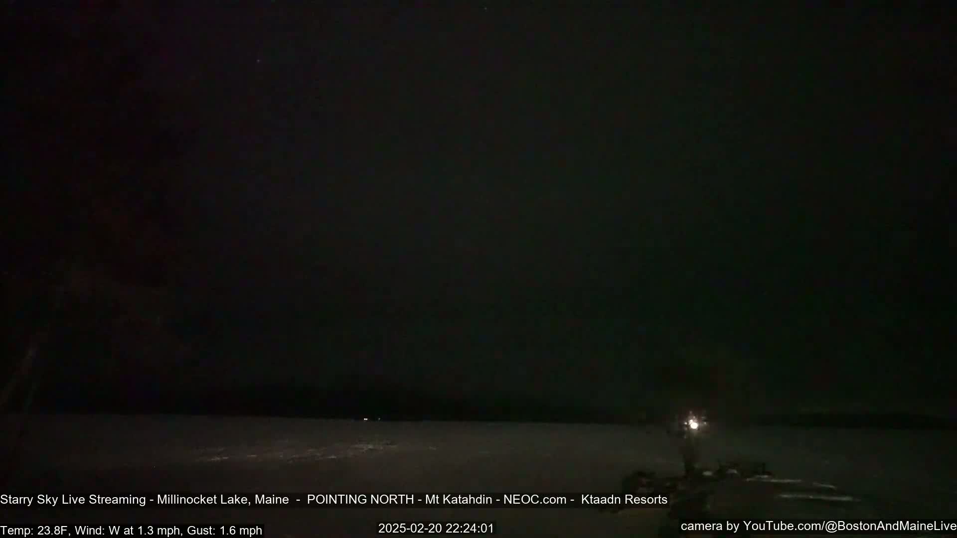 A nighttime view of a dark, snowy lake under a clear, starry sky with a faint light source visible in the bottom right corner.