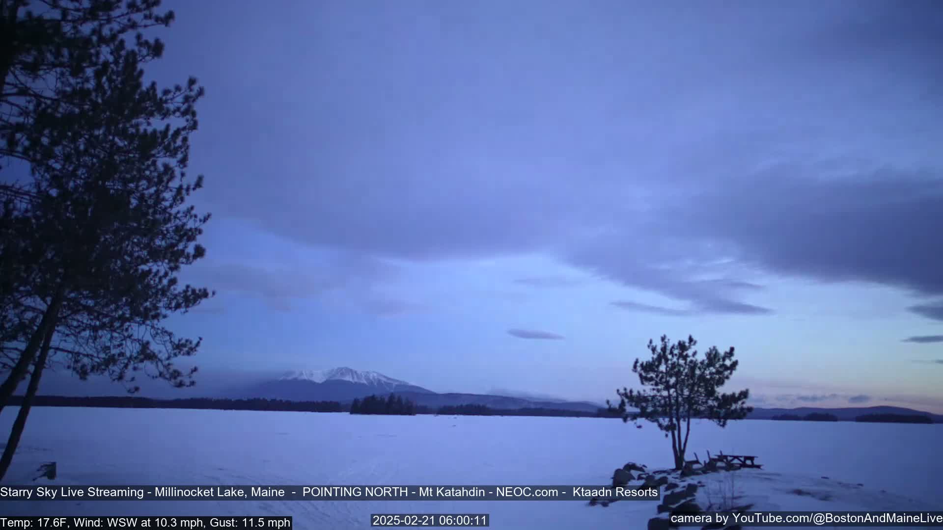A snow-covered lake stretches to a mountain range under a twilight sky with a few clouds and a light wind.