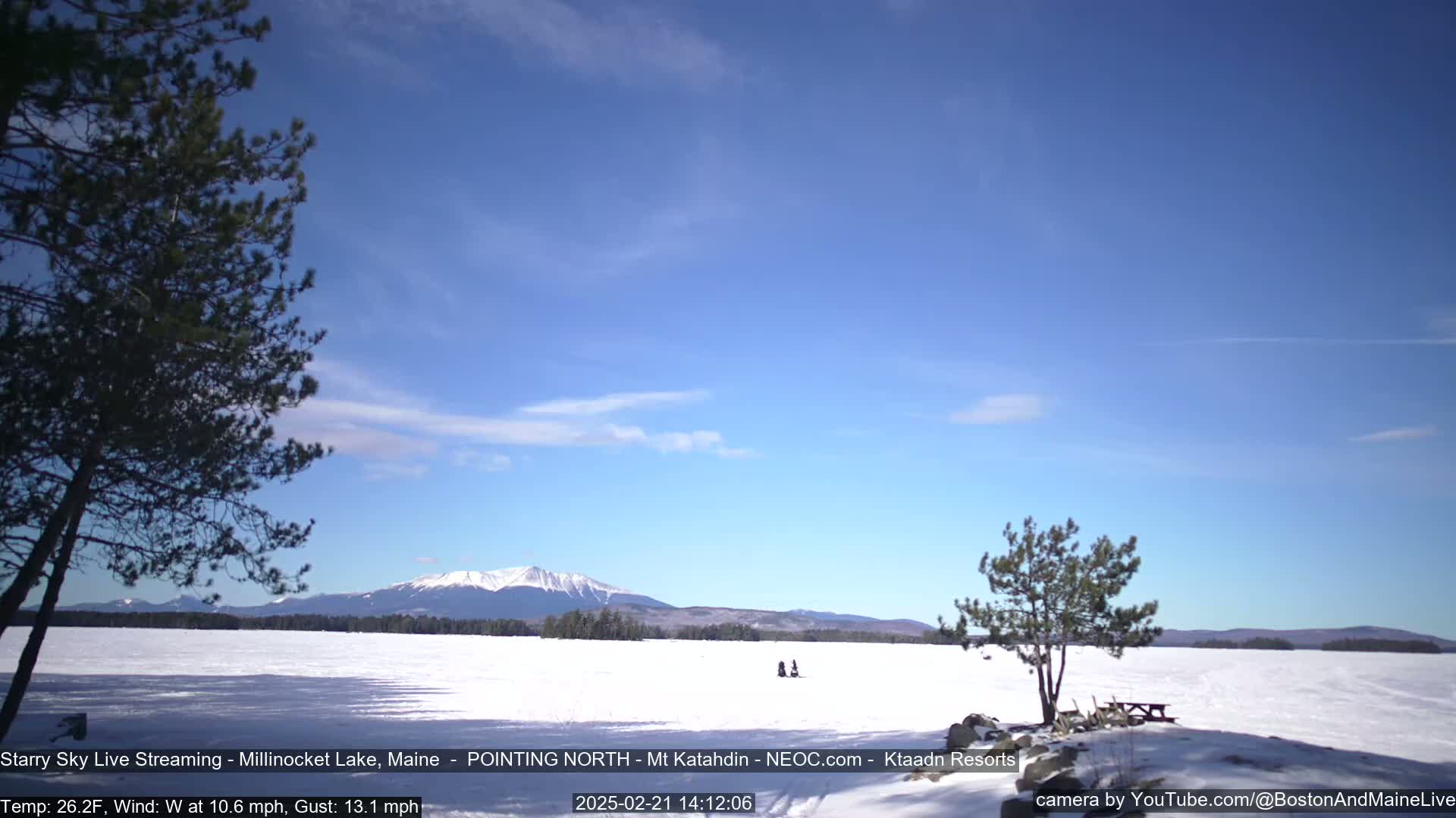 A snow-covered lake stretches out before a distant mountain range under a mostly clear, bright blue sky, with a few wispy clouds and some scattered trees along the shoreline.