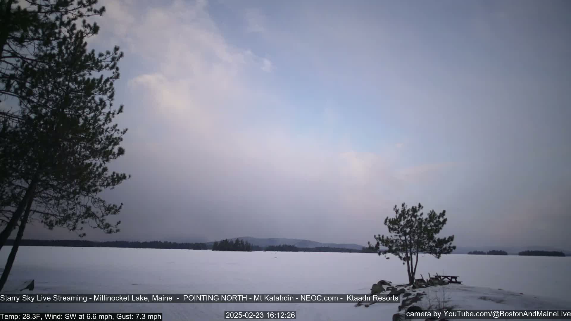 A snow-covered lake is visible under a mostly cloudy, dusky sky, with a few trees silhouetted in the foreground.