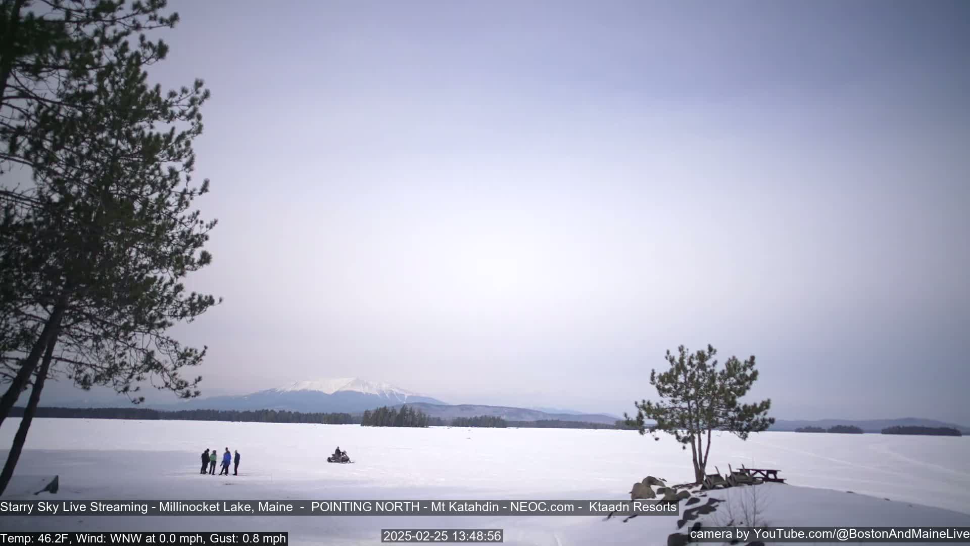 A snow-covered lake stretches to a distant mountain range under a pale sky, with a few people and a snowmobile visible on the ice and some small evergreens in the foreground.
