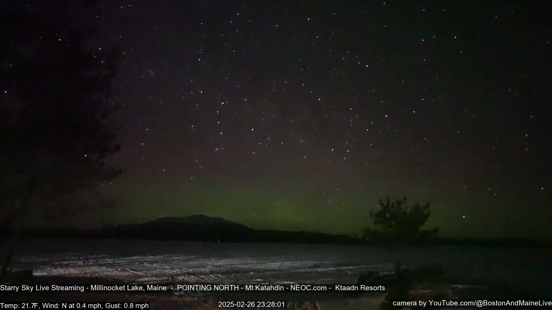 A nighttime long exposure shows a starry sky with a faint aurora borealis over a frozen lake and distant mountains under a clear, cold sky.