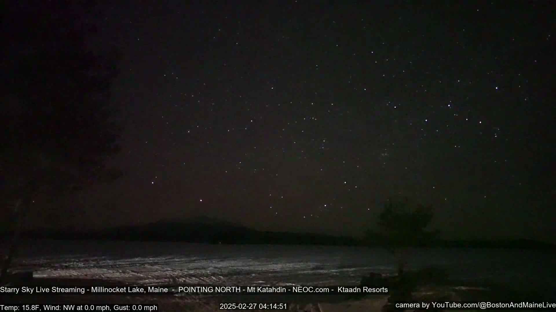 A still, clear night with a starry sky over a snow-covered lake and distant mountains.