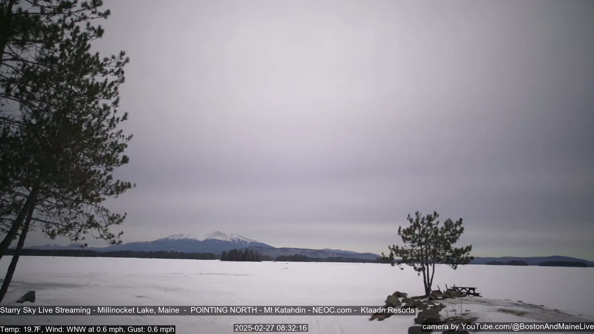 A snow-covered lake stretches to a distant mountain range under a cloudy, overcast sky.