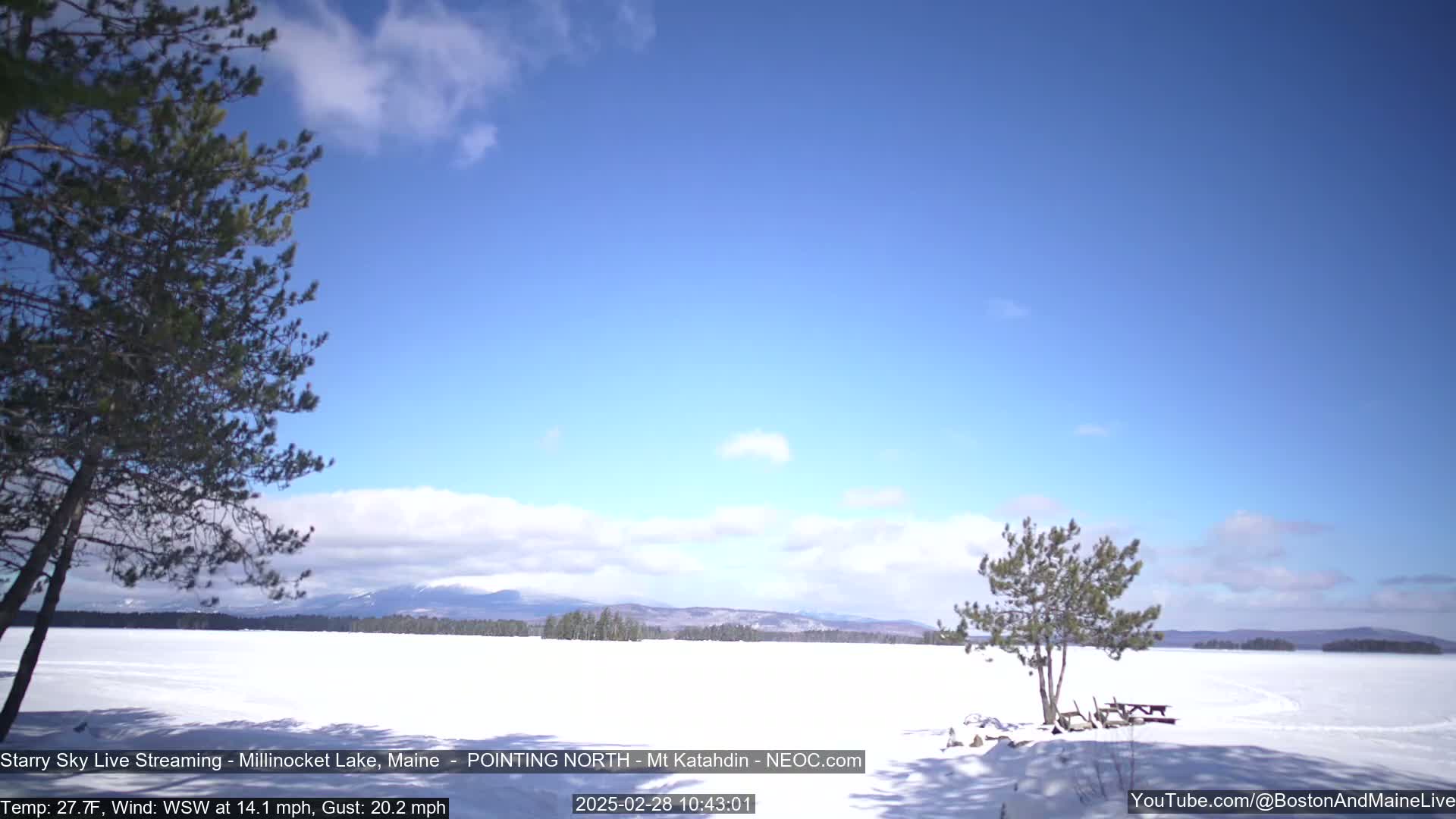 A snow-covered lake stretches to a distant mountain range under a mostly clear, bright blue sky.