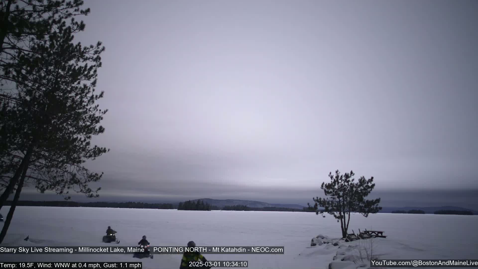 Two snowmobiles travel across a frozen lake under a cloudy, overcast sky.
