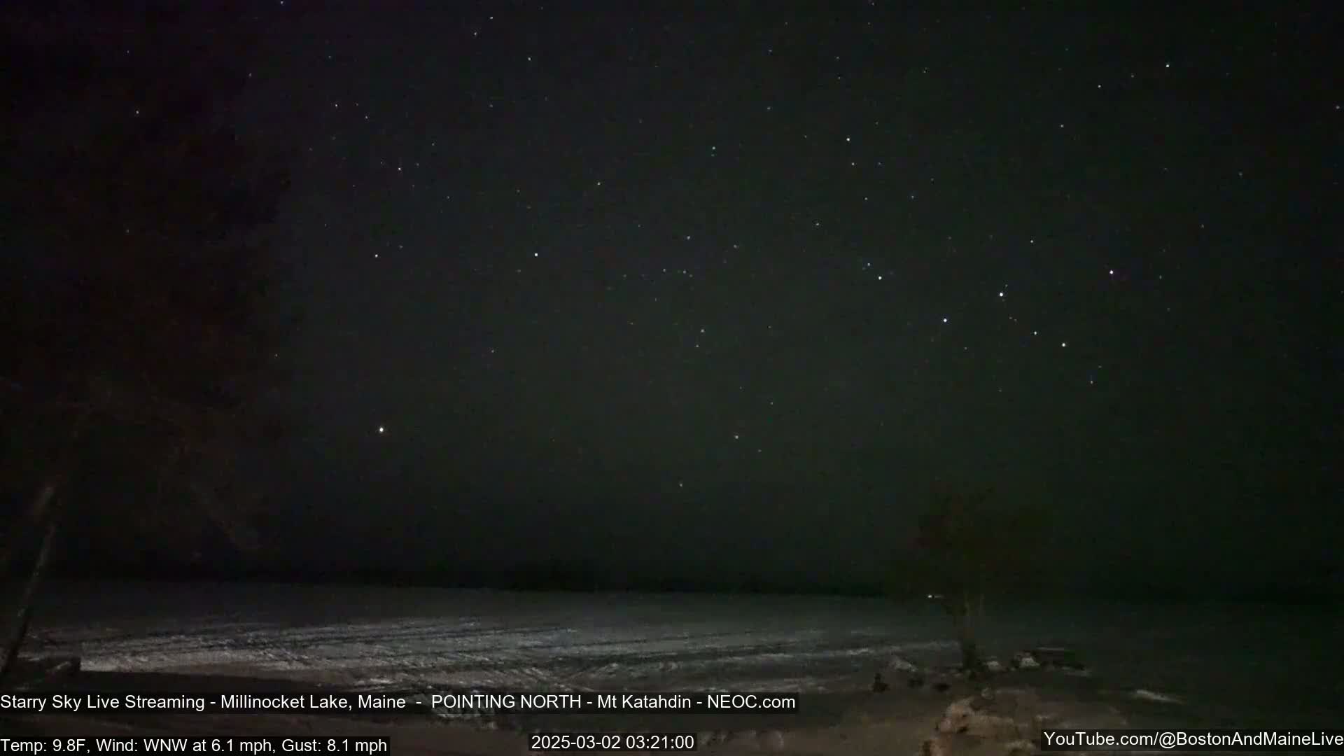 A clear, starry night sky over a dark, snow-covered lake, with a light wind blowing.