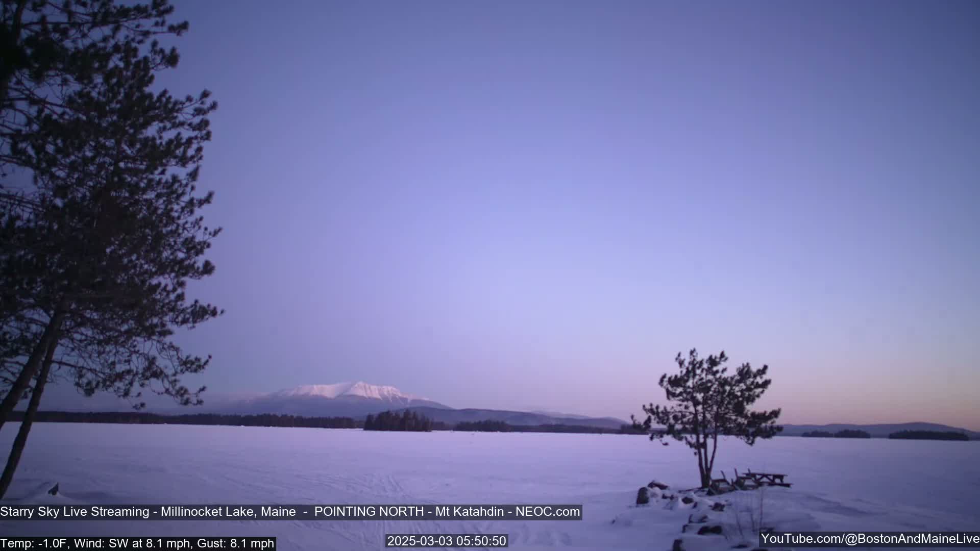 A snow-covered lake stretches to a mountain range under a twilight sky with a light southwest wind.