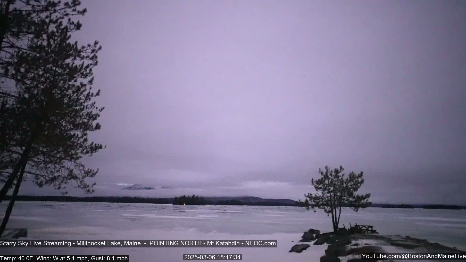 A partly cloudy, cool evening scene shows a frozen lake with two pine trees in the foreground and distant, snow-covered mountains under a pale purple sky.