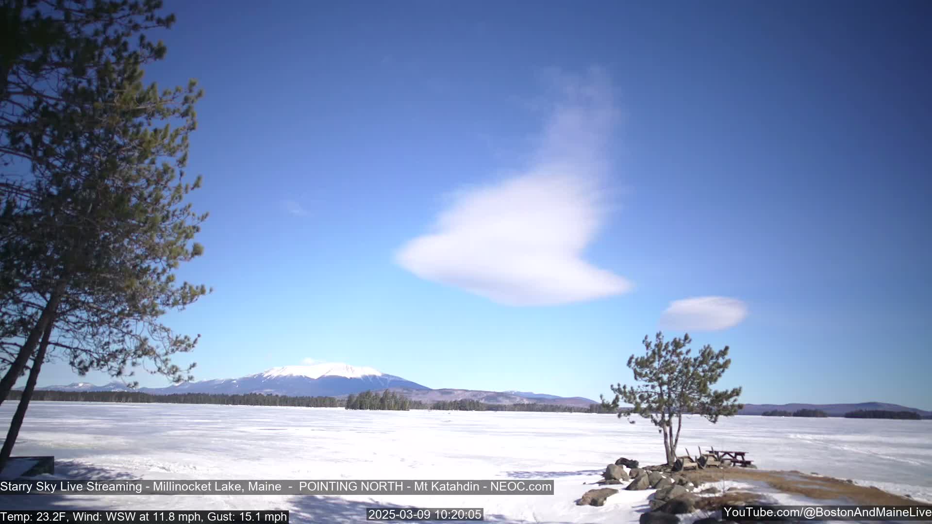 A snow-covered lake stretches to a mountain range under a mostly clear blue sky with a few lenticular clouds, and a light wind is blowing.