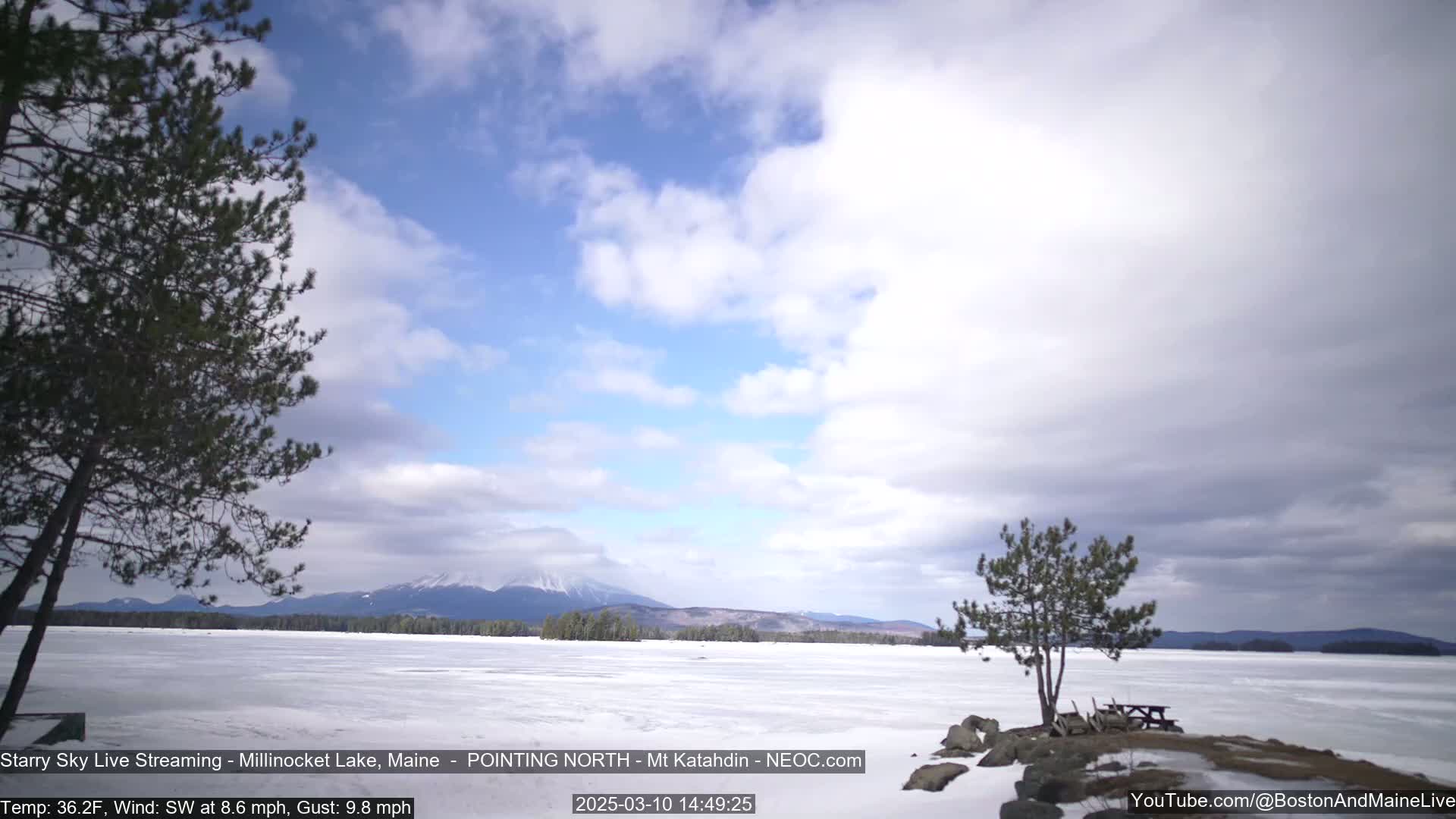 A partly cloudy sky overlooks a frozen lake with a distant mountain range under a partly cloudy sky, with a few trees and a picnic table visible in the foreground.