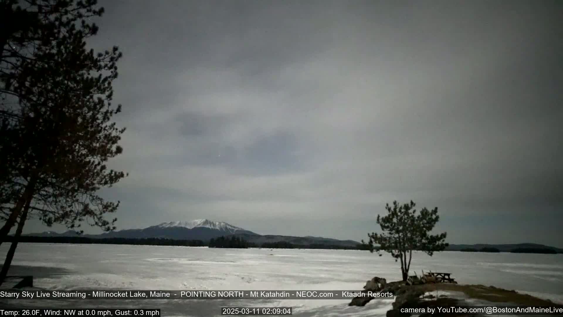 A snow-covered lake stretches to a mountain range under a cloudy, gray sky.