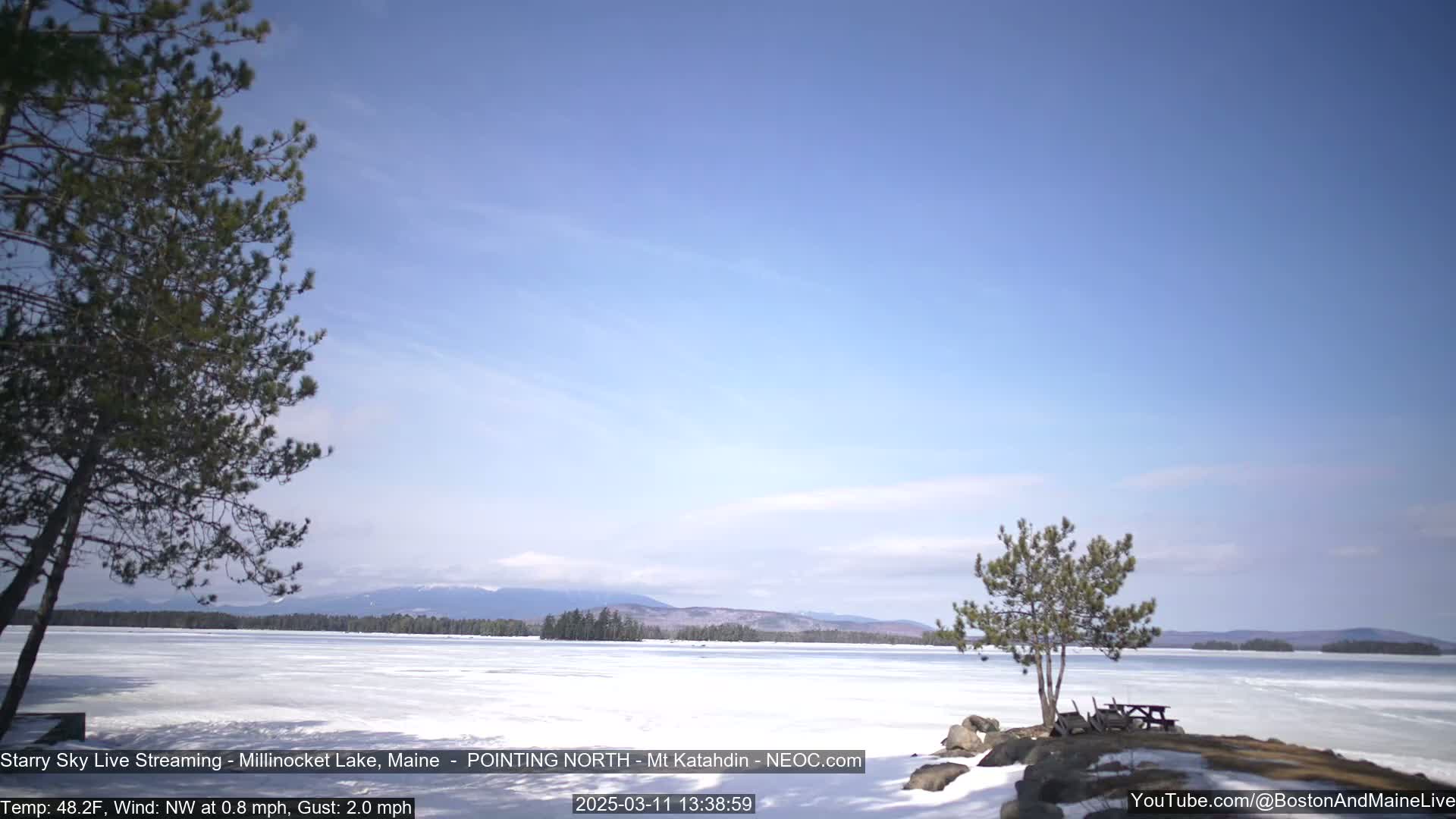 A mostly clear blue sky overlooks a frozen lake with snow-covered banks, a few small trees, and distant mountains.