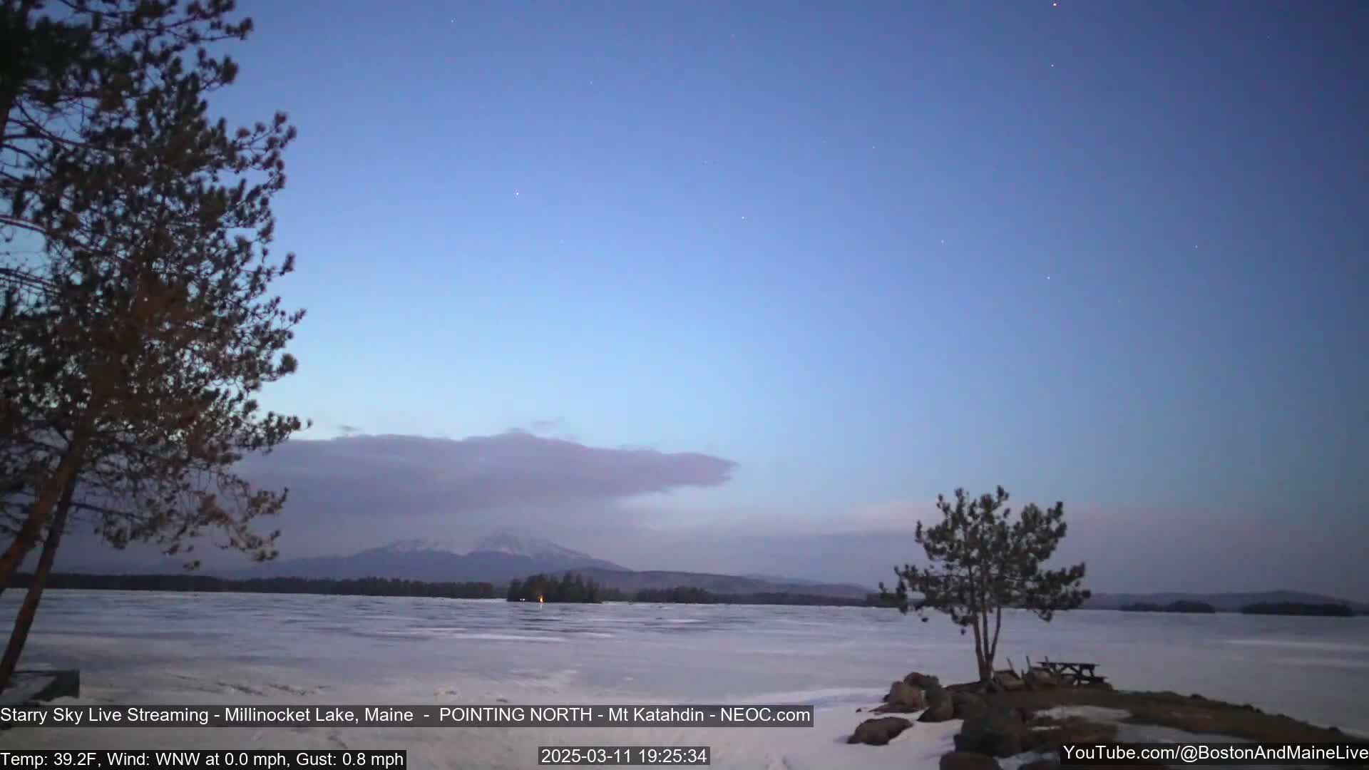 A mostly clear, twilight sky over a frozen lake with distant mountains and a few silhouetted trees in the foreground.