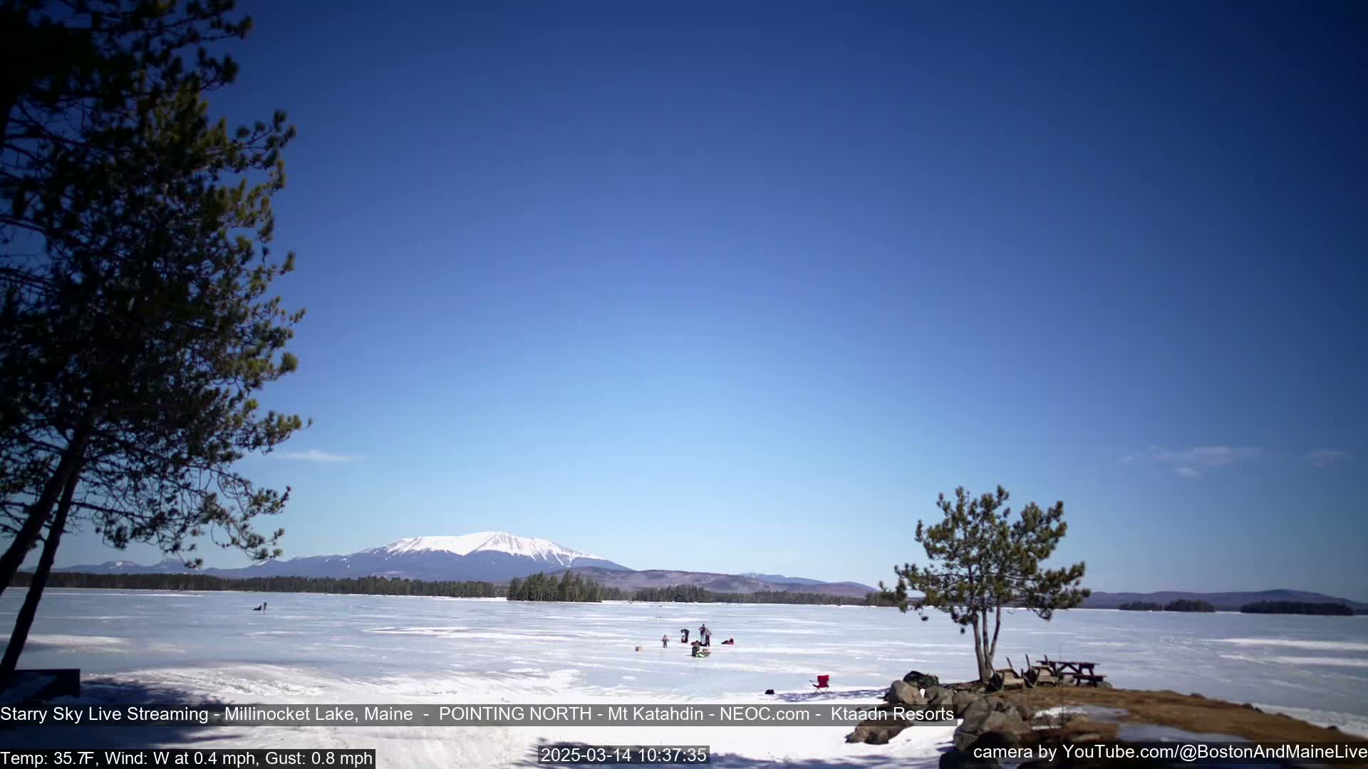 A frozen lake, under a clear blue sky, features several people ice fishing in the distance with mountains visible on the horizon.