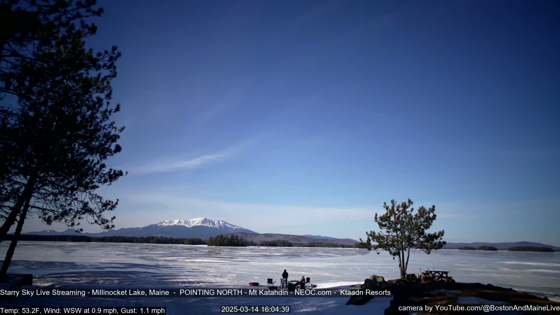 A mostly clear blue sky overlooks a frozen lake with a distant mountain range, and a few people are gathered near the shore.