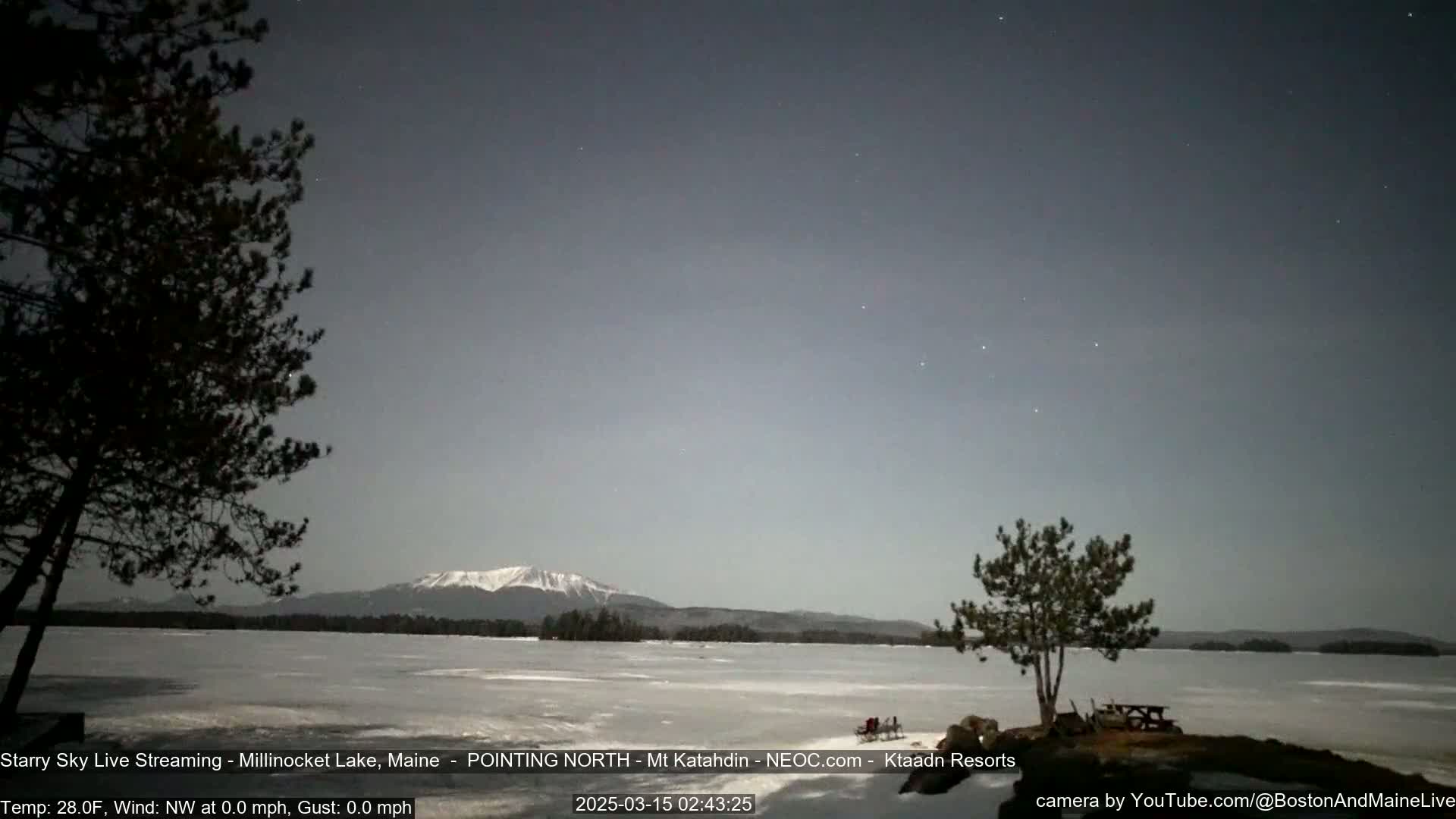 A snow-covered lake under a starry, twilight sky with a mountain range in the distance and silhouetted trees in the foreground.