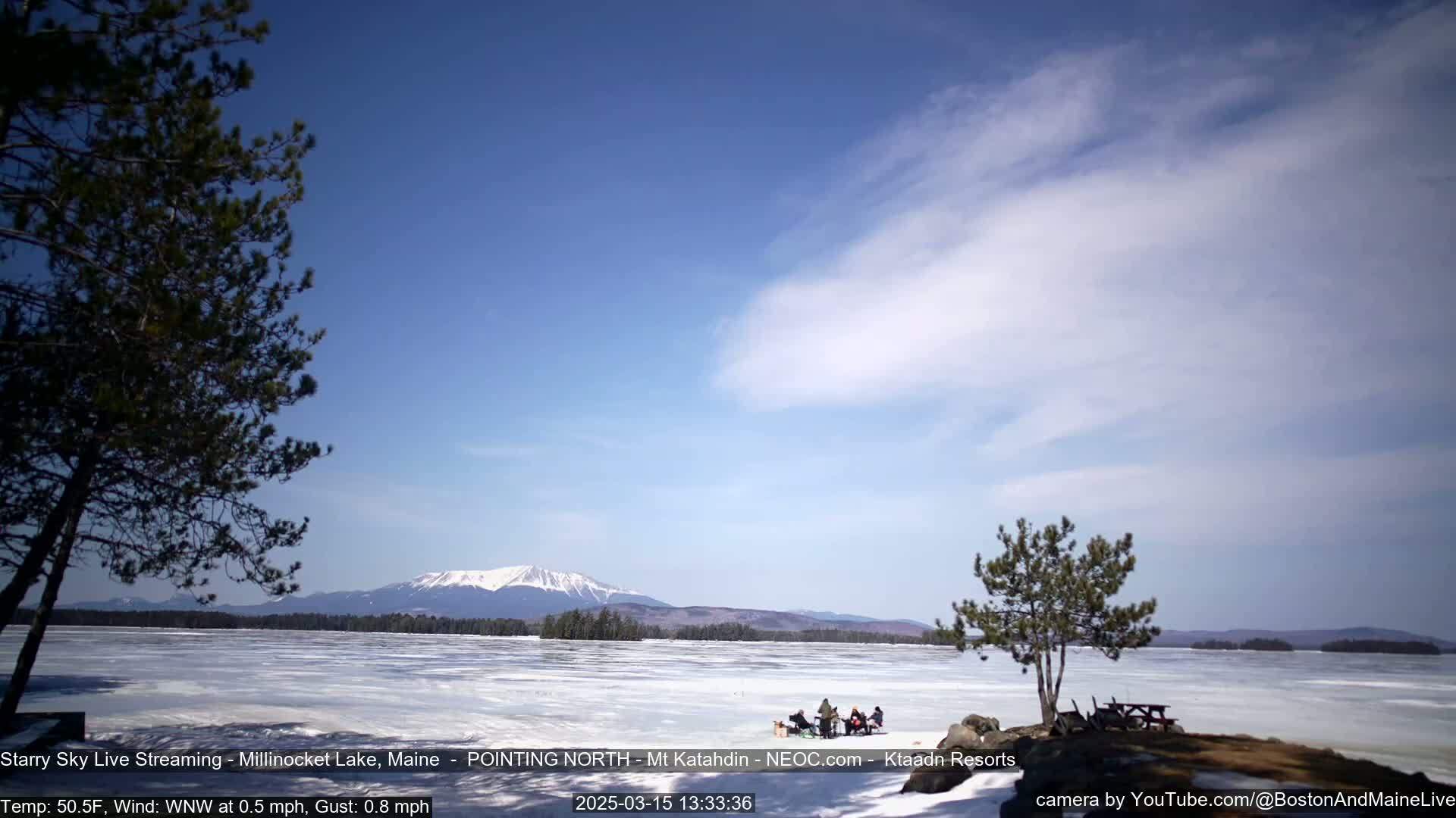 A partially frozen lake stretches toward a snow-capped mountain under a mostly clear, bright blue sky with wispy clouds.