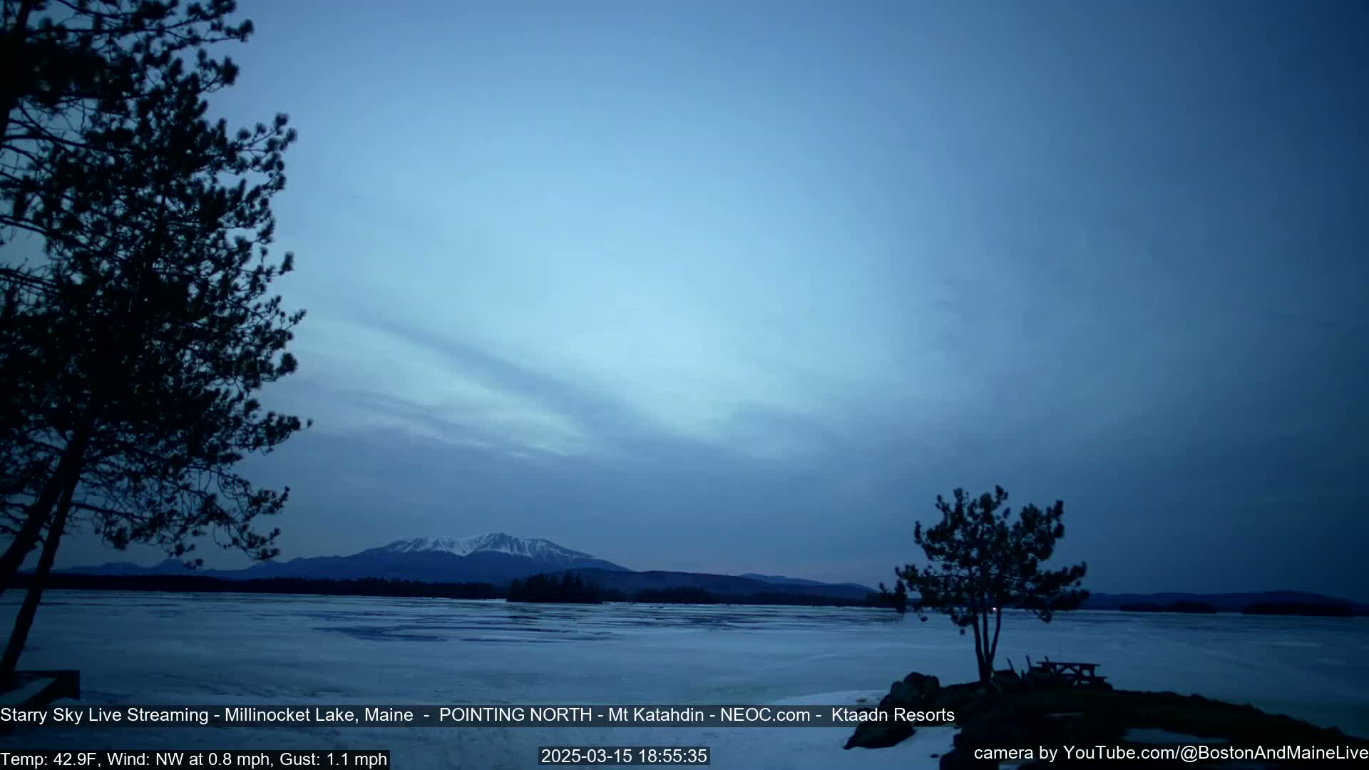 A partly cloudy, twilight scene shows a partially frozen lake with a distant mountain range under a dusky blue sky, framed by silhouetted trees.