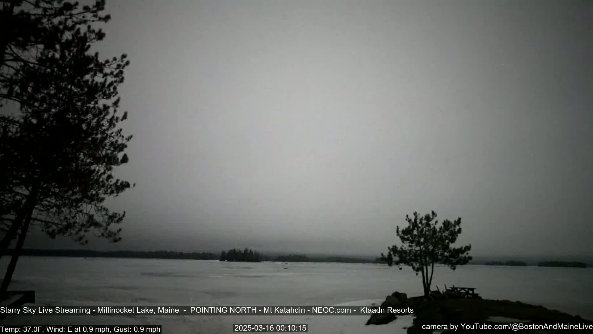 A mostly overcast, gray day shows a partially frozen lake with dark trees on either side of the shore.