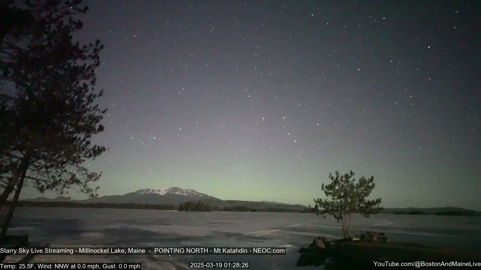 A starry night sky over a frozen lake, with a mountain range in the distance and silhouetted trees in the foreground, under calm, cold conditions.