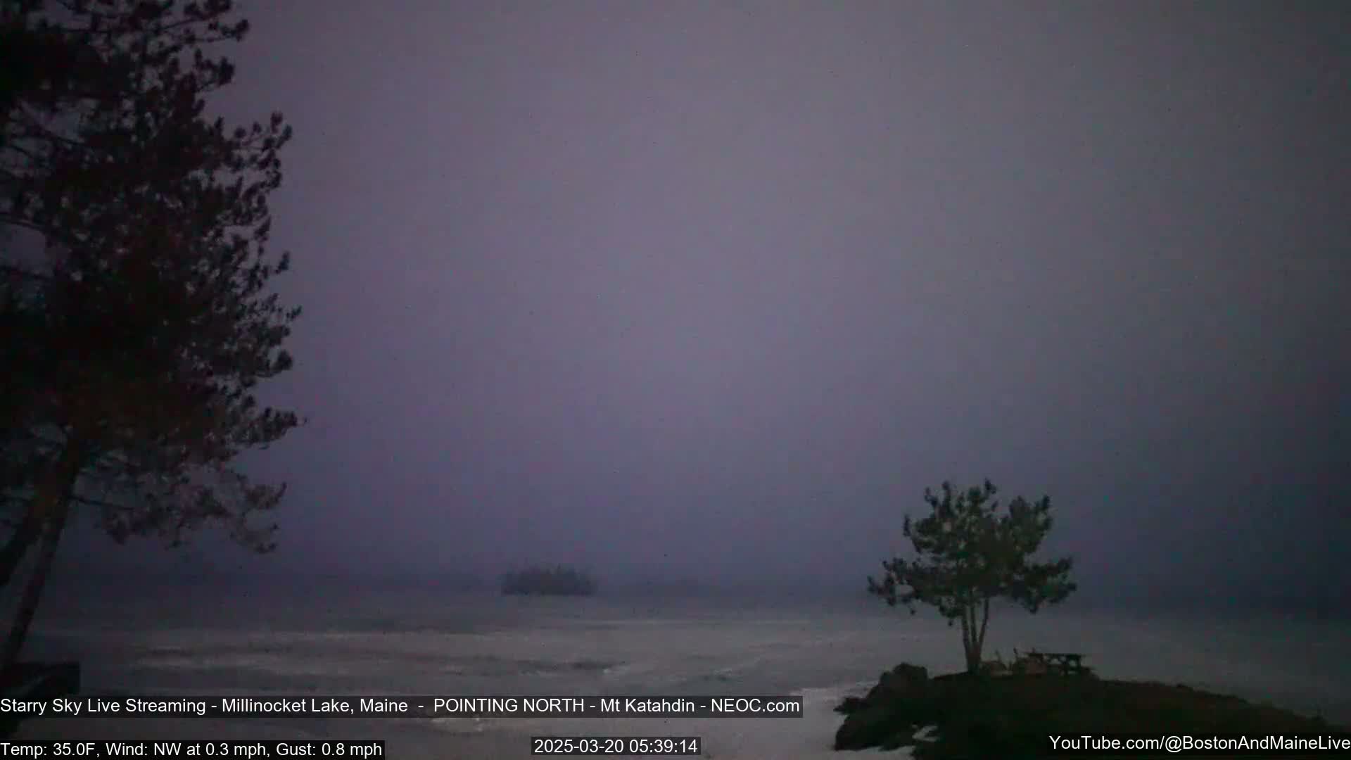A foggy, dark gray morning scene shows a lake with a small island in the distance and silhouetted trees on either side of the foreground.